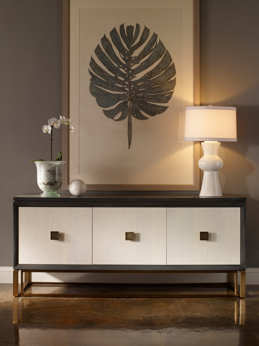 Wallace Storage Console cabinet in black and white colors with three doors and three adjustable shelves, satin brass plated base and hardware. Presented in front of a wall with an art above it and a lamp and vase on it.