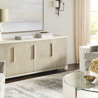 Axis Media Console cabinet in light colors with four doors, two adjustable shelves, one interior drawer, and standard bronze hardware. Placed in a modern living room with books and a lamp on it.