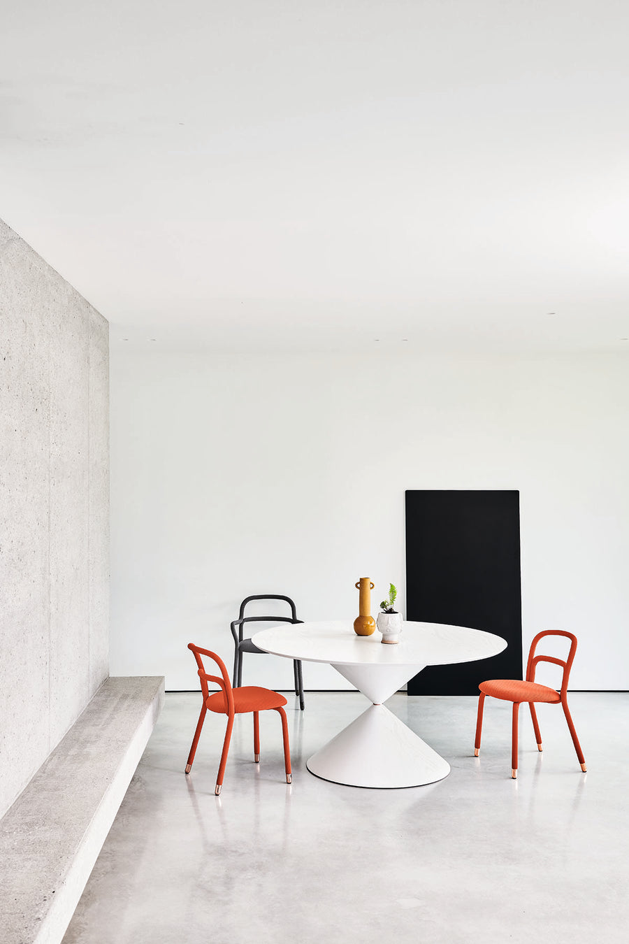 Fixed round Clessidra Dining Table with white base and white top. Placed in a white room with two orange chairs and one black chair.