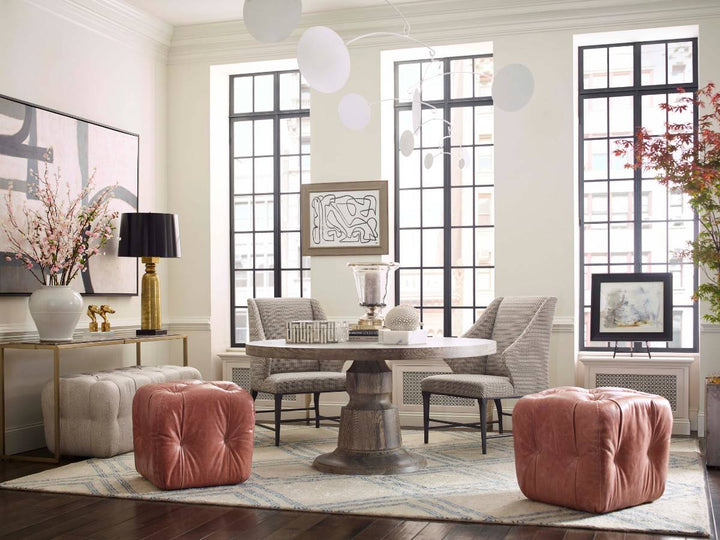 A stylish dining room with a round pedestal table, upholstered chairs, leather ottomans, and statement artwork, featuring natural light and bold accents.