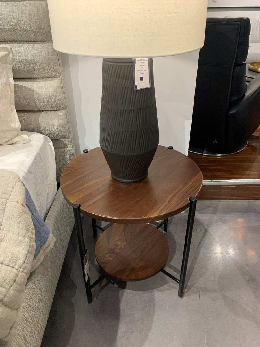 Manhattan Side Table with the racetrack oval, asymmetrical silhouette, and unique joinery between metal, glass and wood pieces. Placed besides a bed and with a lamp on it.