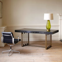 Three drawer Marcel Desk in Variegated Black with brass wrap placed in a room with a matching chair and a lamp on top of it.