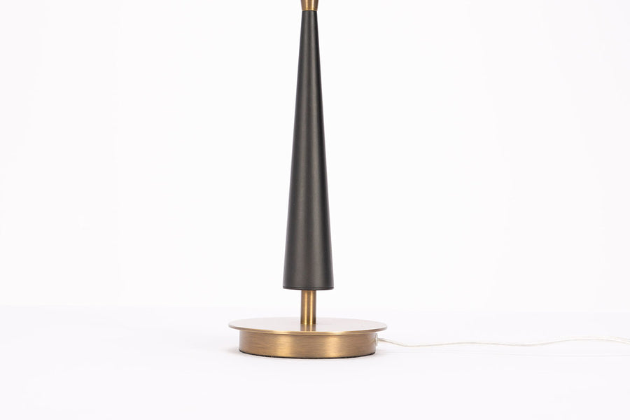 Morrison Lamp with a round Antique Brass base, white drum shade and candlestick silhouette body that combines Antique Brass with Matte Black. Closed up view on body and base.