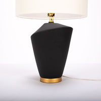 Gavin table lamp with a drum white shade and modern black ceramic body in an unique asymmetric design and a contrasting gold base. Closed up view on the body and the base.