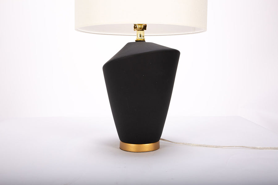 Gavin table lamp with a drum white shade and modern black ceramic body in an unique asymmetric design and a contrasting gold base. Closed up view on the body and the base.